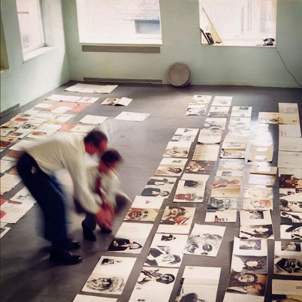 Pagine di rivista disposte in file sul pavimento di uno studio, due figure mosse chine a riordinarle