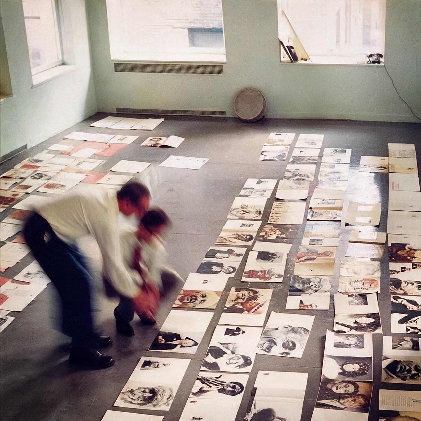 Pagine di rivista disposte in file sul pavimento di uno studio, due figure mosse chine a riordinarle