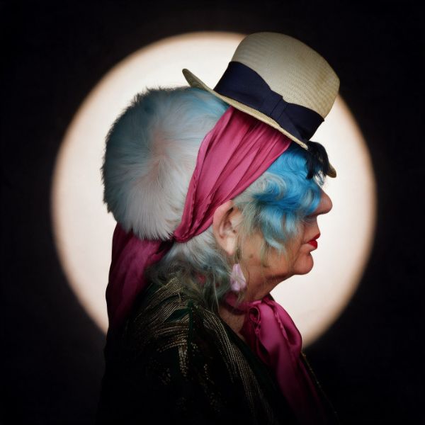 Di profilo, una donna dai capelli azzurri con un cappello di paglia dal nastro blu, una pelliccia bianca e una sciarpa di seta fucsia, davanti a un cerchio di luce su fondo nero