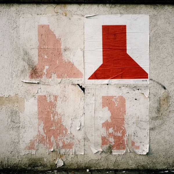 Quattro manifesti identici con un segno rosso affissi su un muro: tre consumati dal tempo, uno intatto